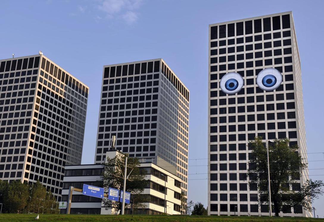 This image depicts three city towers, one of which has two large blue eyes designed onto the face of the building. 